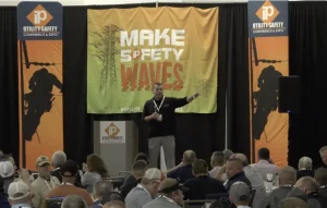 A speaker presents to an audience in front of a banner that reads "Make Safety Waves" at the iP Utility Safety Conference & Expo.